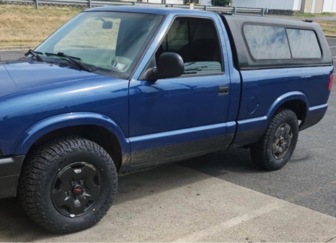Blue pickup truck with a black camper shell and black wheels parked on a paved lot.