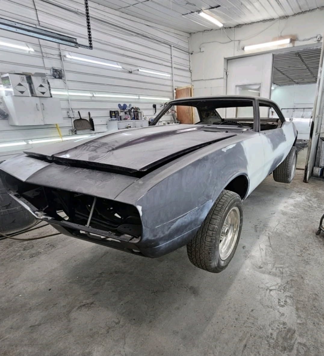 A classic muscle car in primer grey sits partially restored in an auto body workshop.