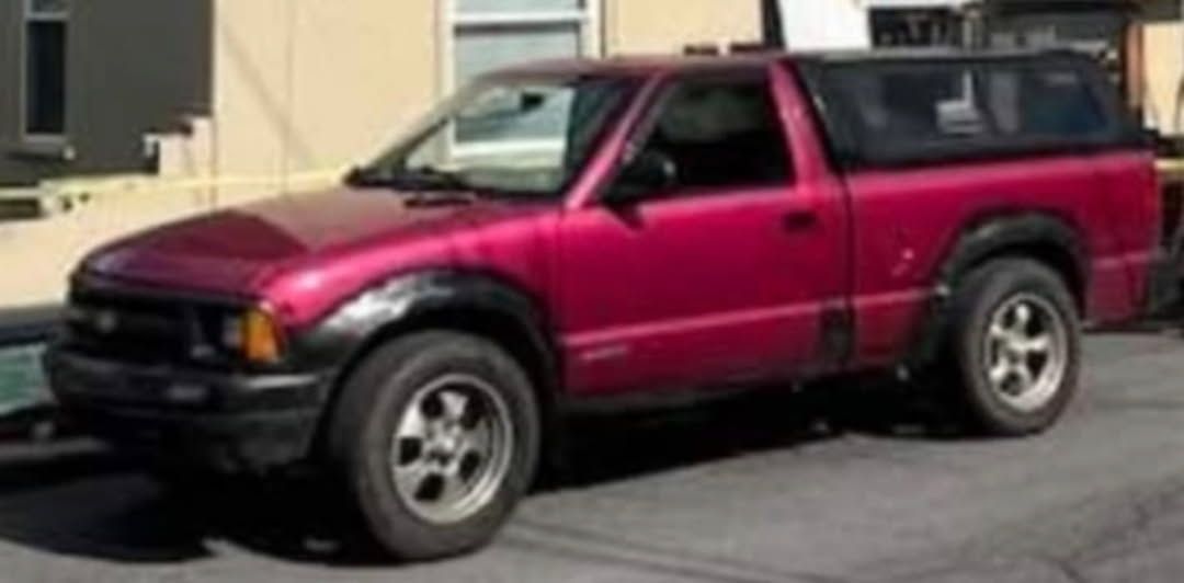 A magenta pickup truck with a black camper shell parked on an asphalt street in front of a building.