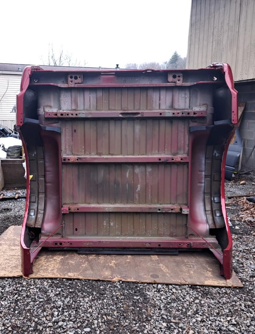 The underside of a maroon truck bed, resting upright on a wooden board against an outdoor, industrial background.