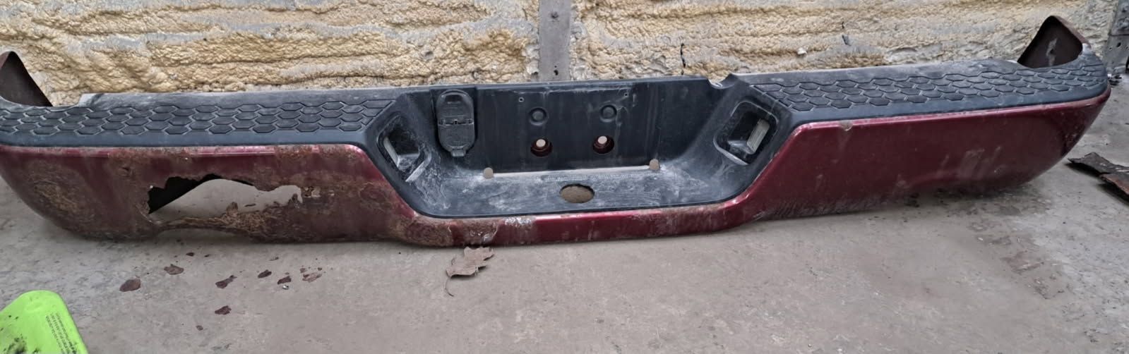 A maroon, damaged truck rear bumper with a black step pad lying on a concrete surface.