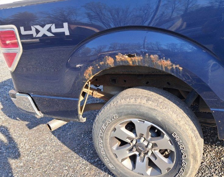 Blue pickup truck with rusted rear wheel well and exposed inner fender, parked outdoors