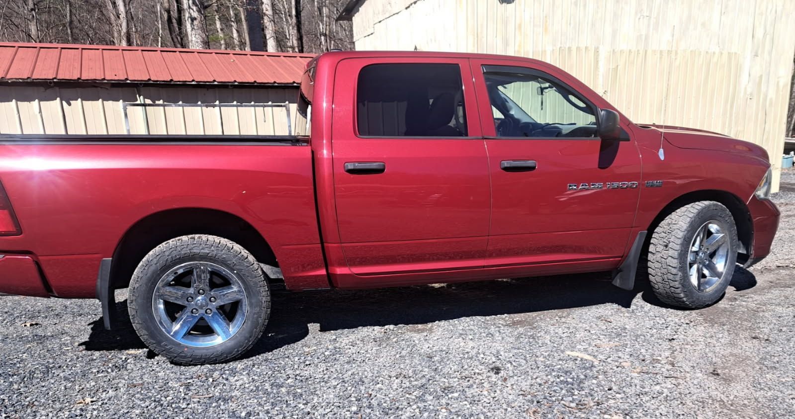 Red pickup truck parked on gravel beside a wooden fence and building.