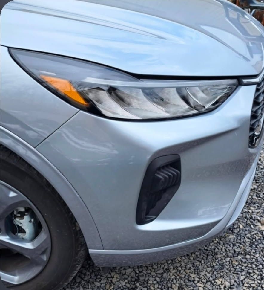 Close-up of a silver car’s front left headlight and bumper on gravel