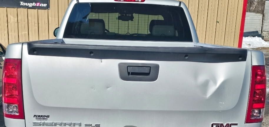 Rear view of a silver GMC pickup with a dented tailgate parked outdoors in front of a wooden fence.