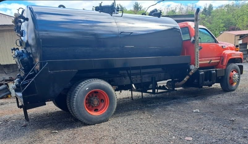 Red dump truck with a large black tanker bed parked on a gravel lot under a cloudy sky