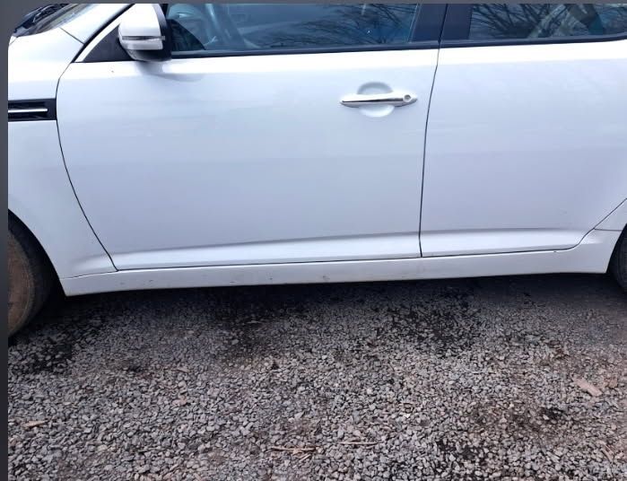 Side view of a white car parked on gravel, showing the front door and side mirror.