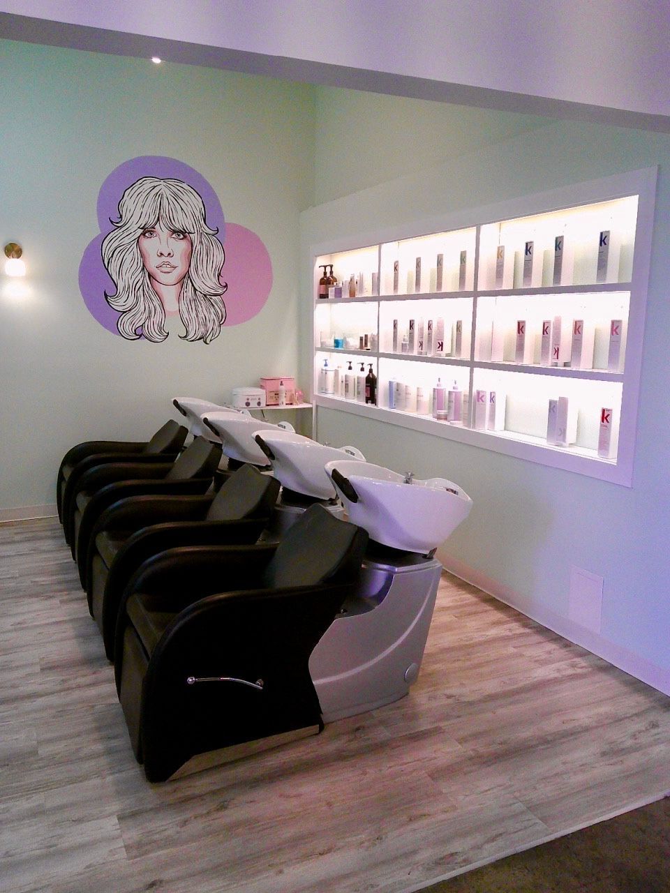 Salon with black and white shampoo chairs beneath a lit product display and a pop-art wall portrait