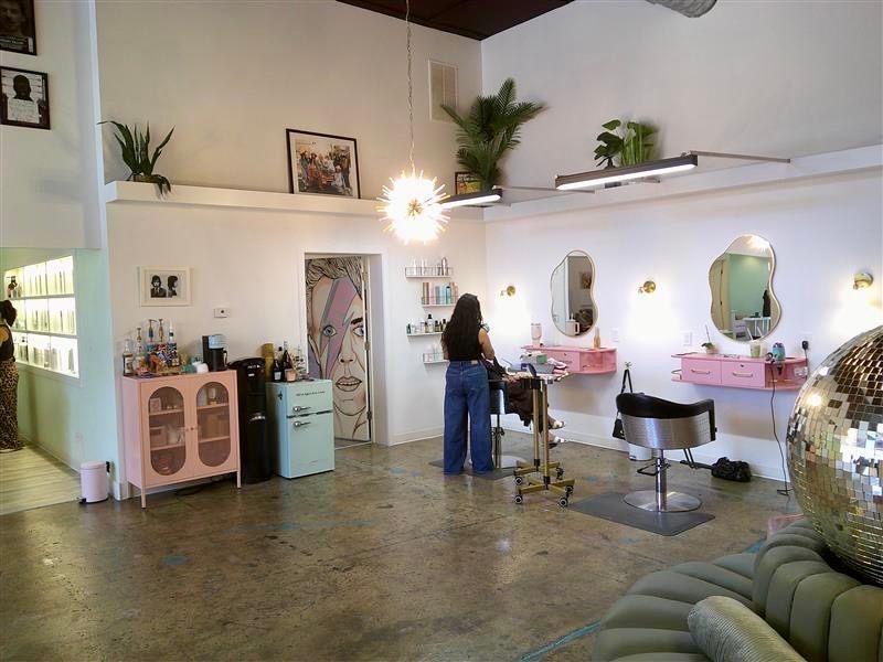 Bright salon interior with styling chairs, mirrored stations, plants, and a person standing by the counter