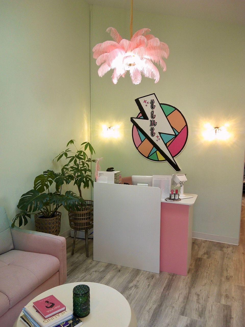 Pink salon reception with modern art, soft lighting, plants, and a white-and-pink counter in a cozy room