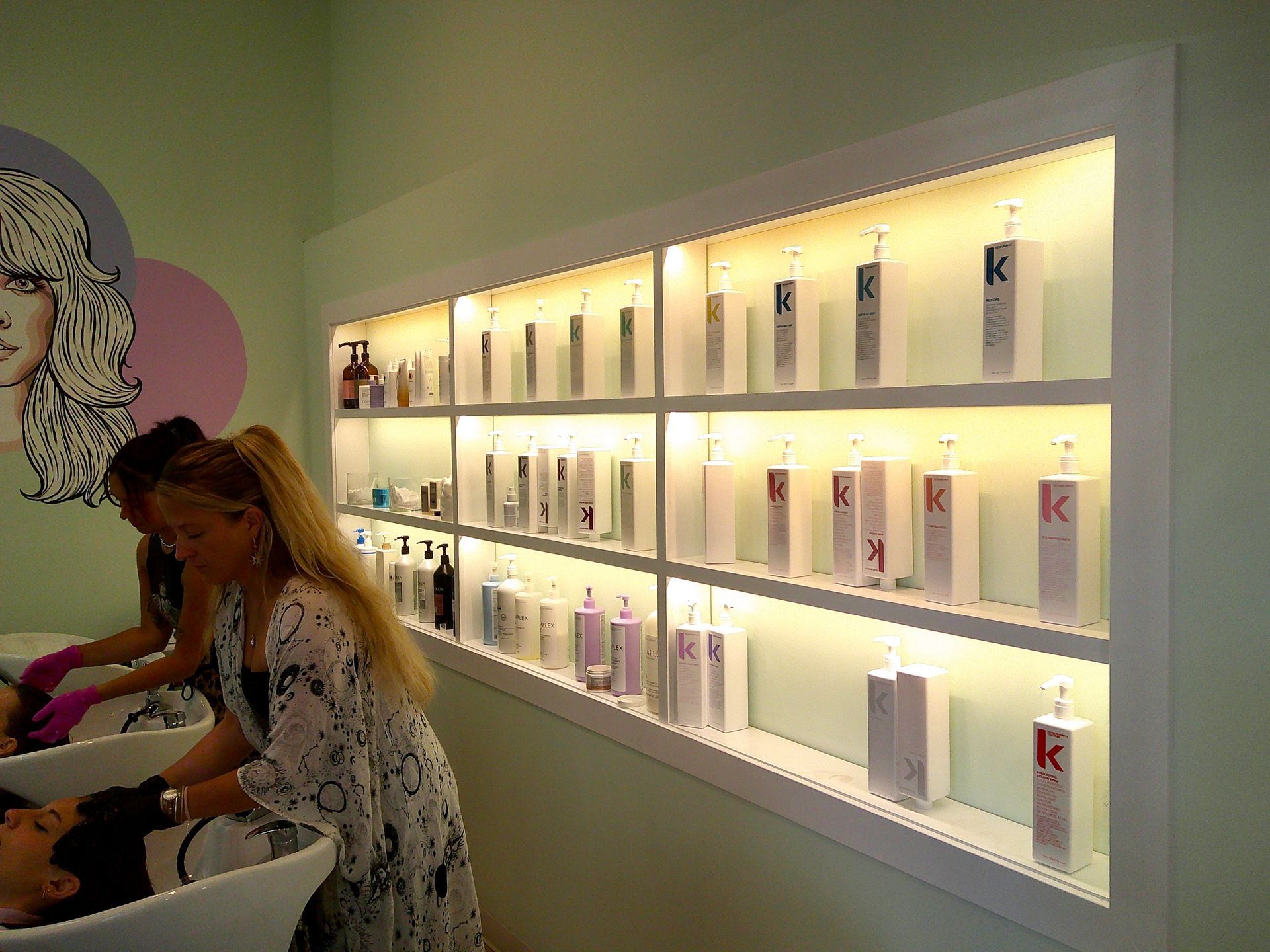 Two people using a nail salon station beside a backlit wall of nail polish bottles