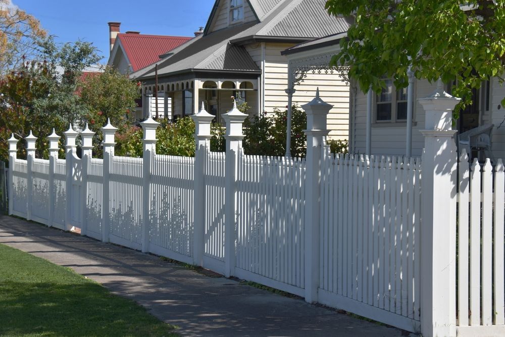 White picket fence in front of light-colored house. Green lawn and tree on left, blue sky above.