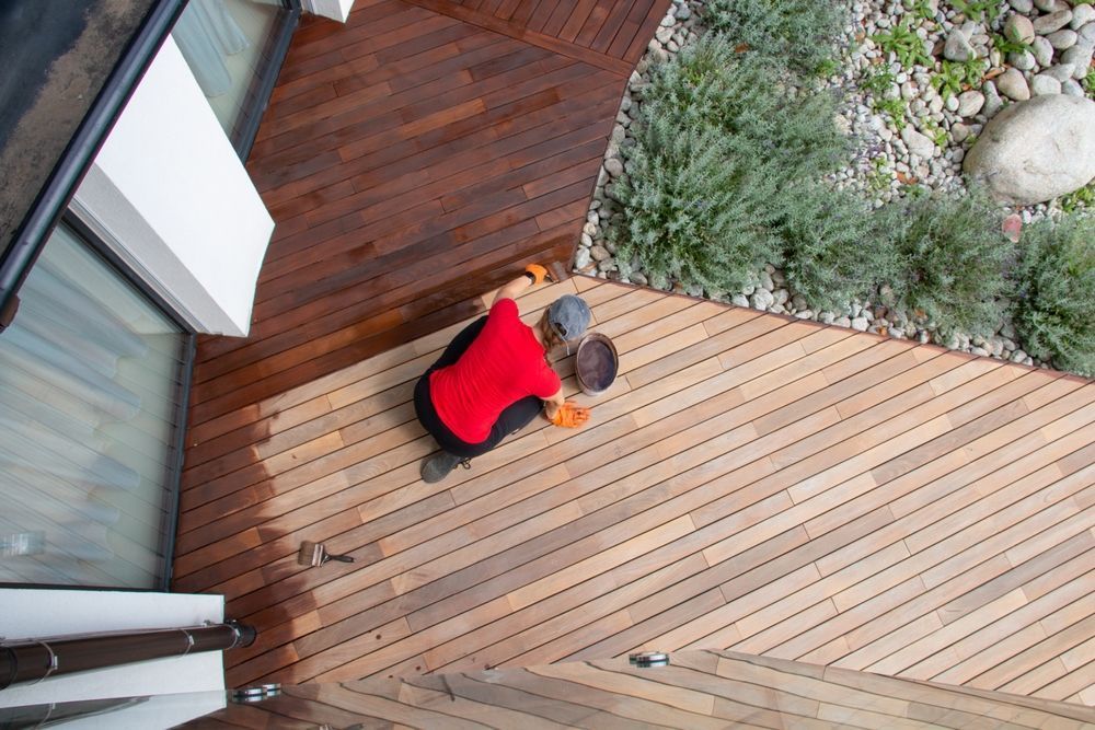 Person staining a wooden deck outdoors, near a window and plants.