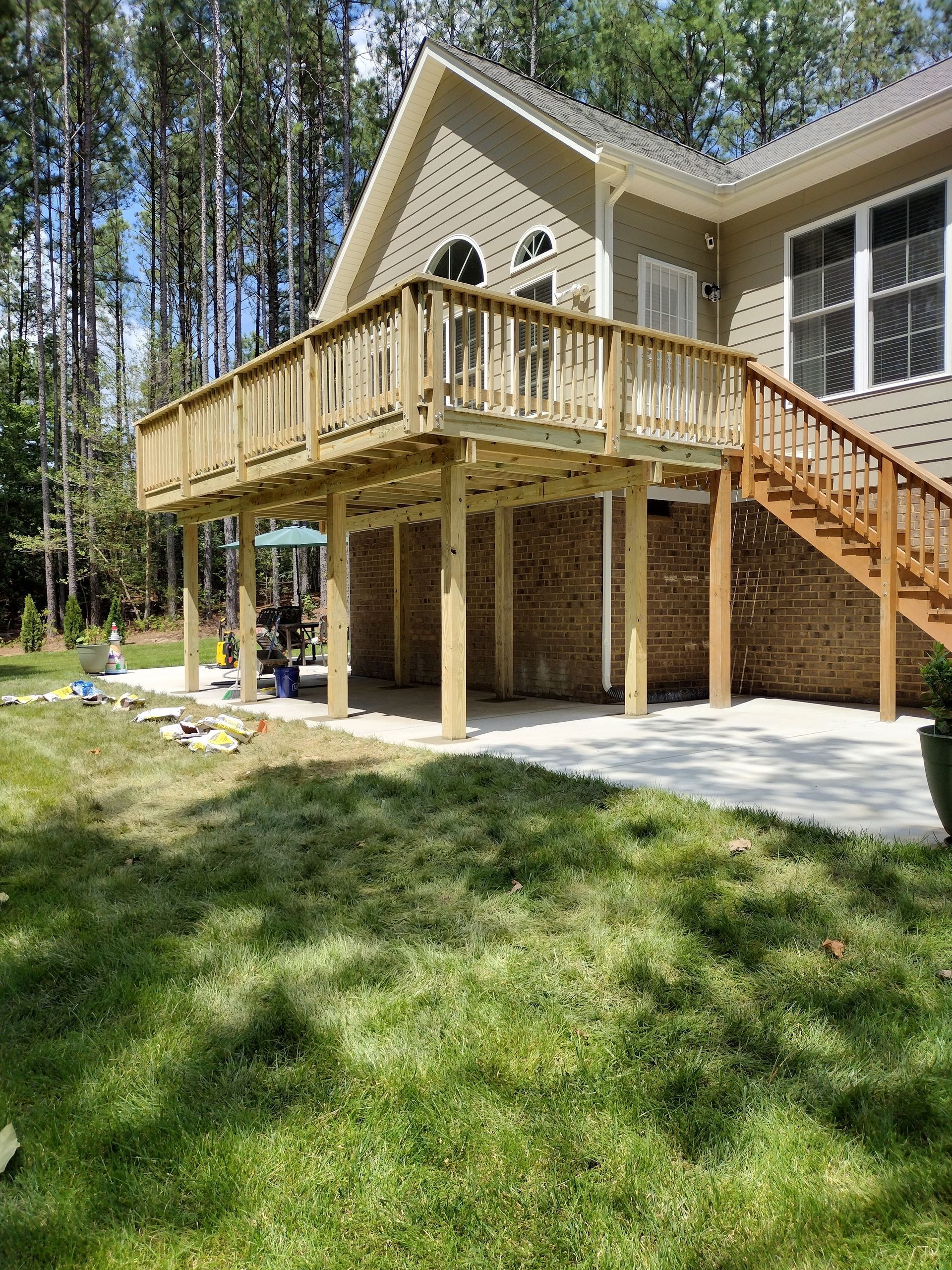 Wooden deck attached to a house with brick foundation and concrete patio below, surrounded by grass and trees.