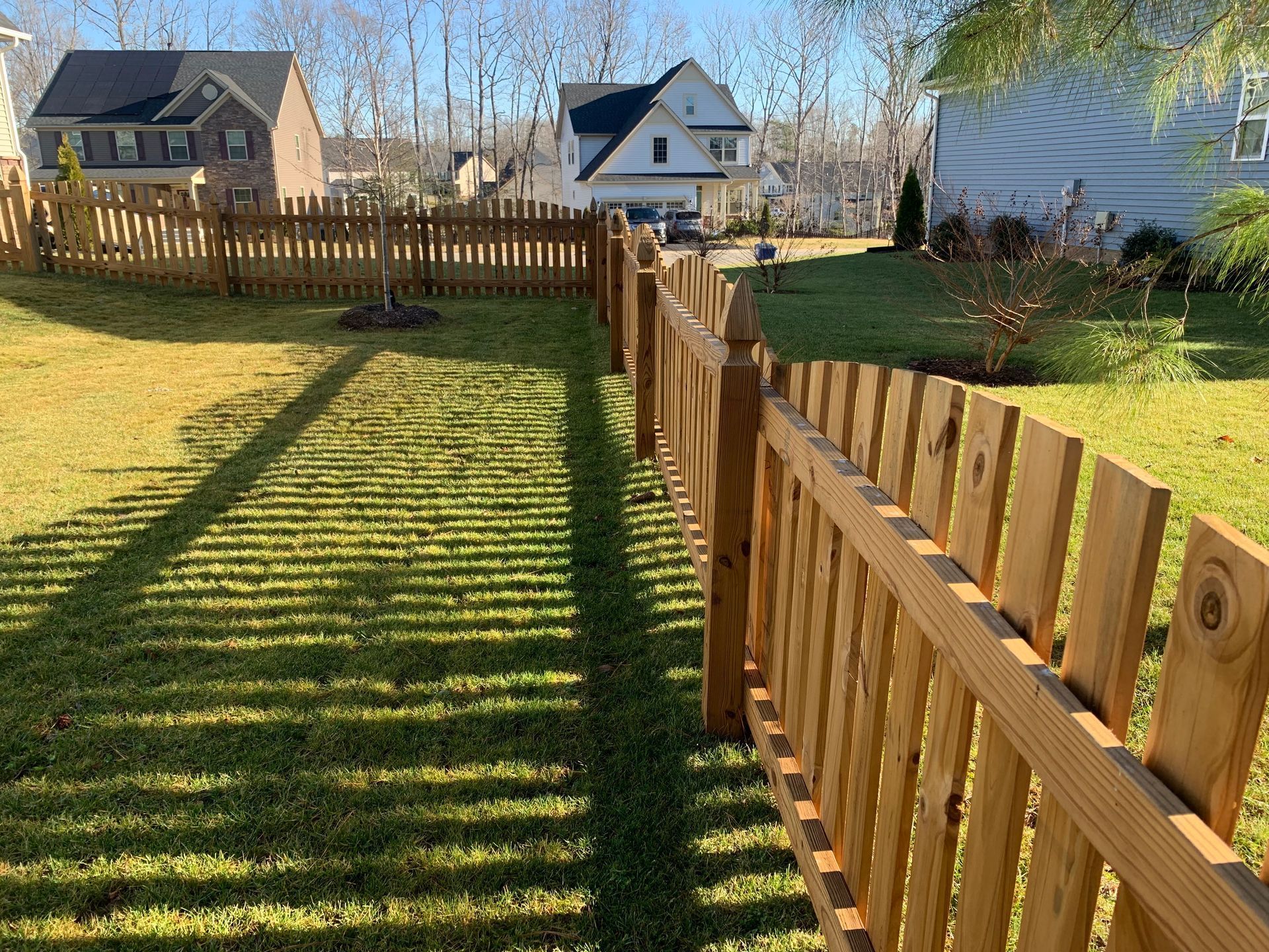 Wooden fence casting long shadows across a grassy yard, with houses in the background on a sunny day.