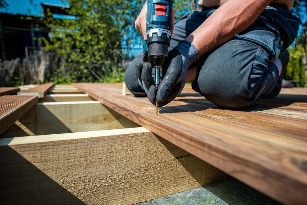 Person kneeling, using a drill to attach wooden deck boards outdoors.
