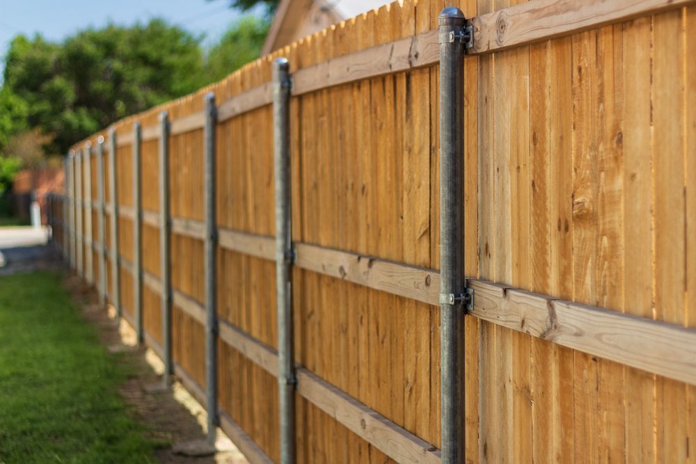 Wooden fence with vertical planks and horizontal rails, supported by metal posts, along a green lawn.