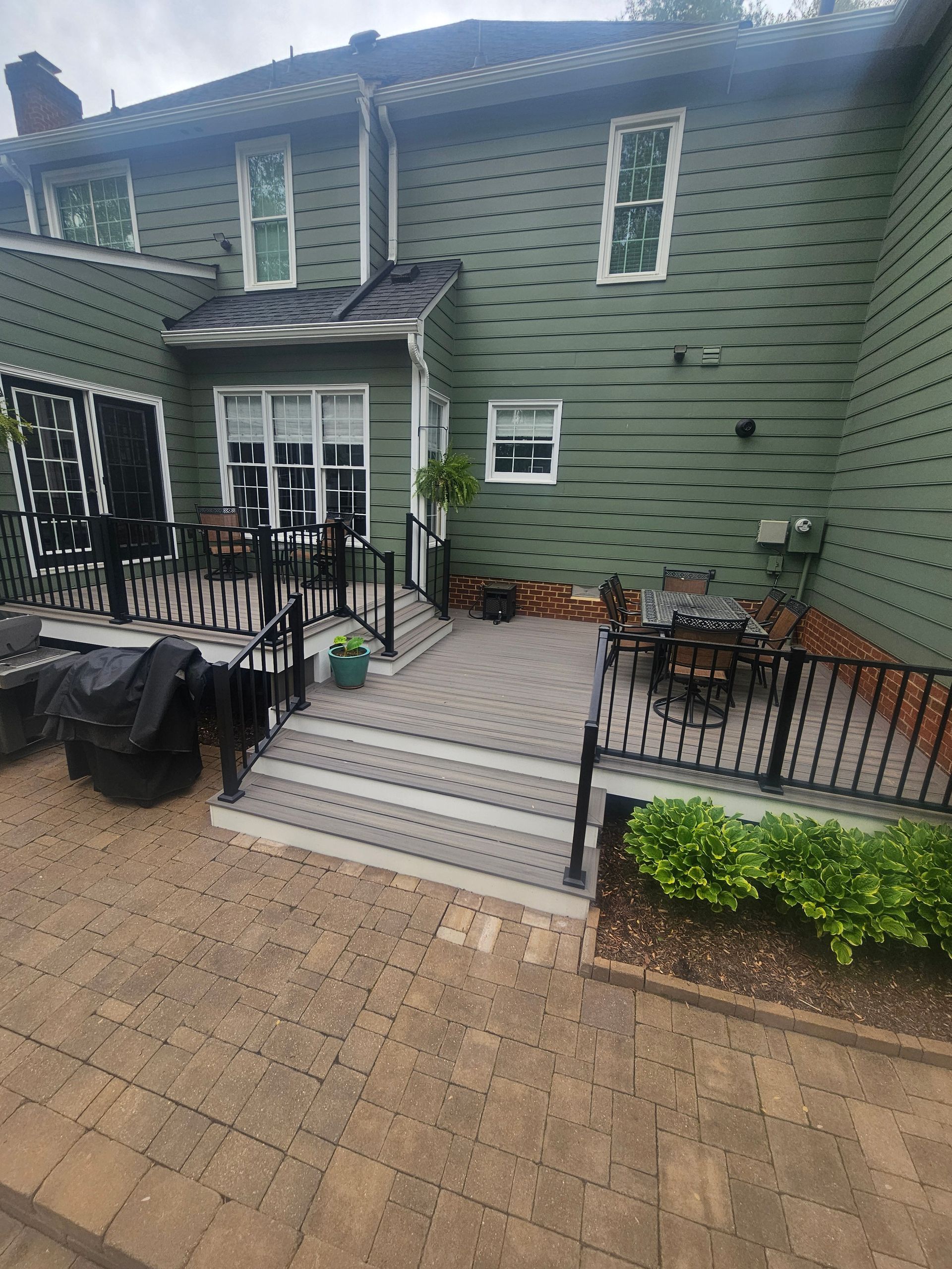 Back of a green house with a multi-level deck, patio, and black railing.