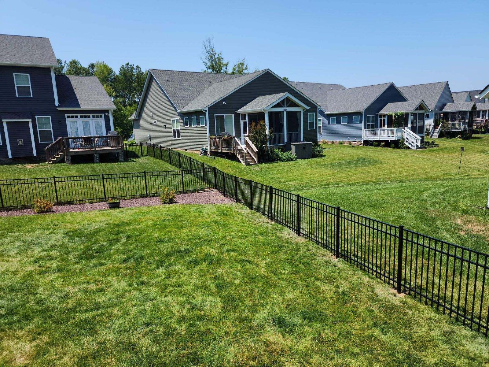 Black metal fence in a grassy yard, with several houses in the background under a blue sky.