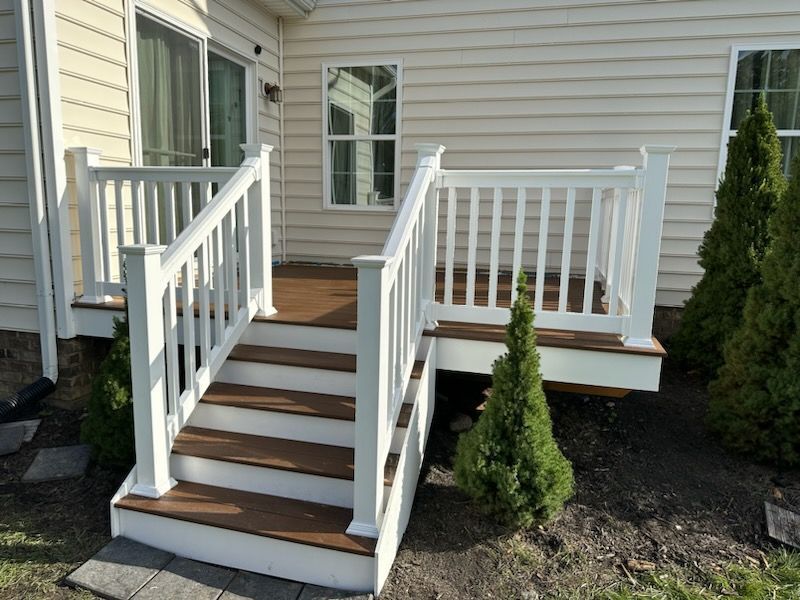 White-railed deck with stairs attached to a light-colored house. Brown deck boards, small evergreen trees.