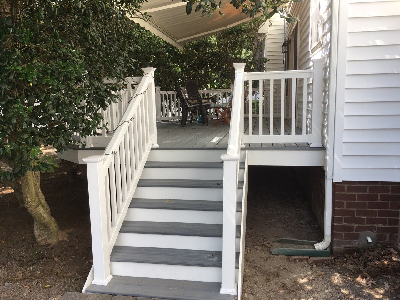 White deck with gray steps, railing, and a view of a seating area, beside a white house with a brick foundation.