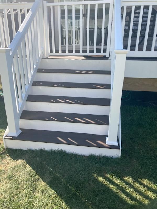 White deck stairs with dark brown treads, leading to a deck. Sunlight casts shadows on grass.