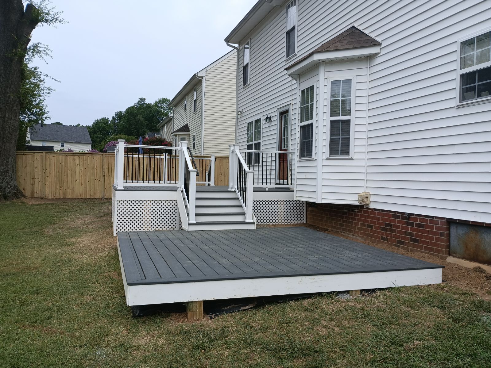 Gray composite deck attached to a white house with a set of stairs. Green grass, wooden fence.