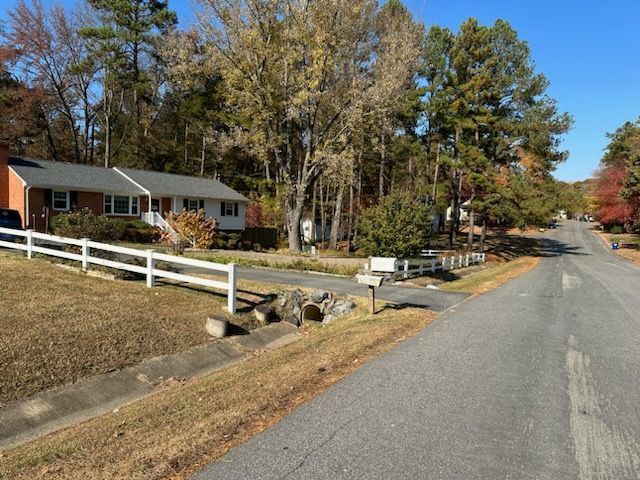 Residential street with houses, white fences, and autumn trees. Paved road curves into the distance.