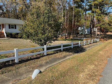 White fence bordering a grassy area next to a road, with houses and trees in the background.