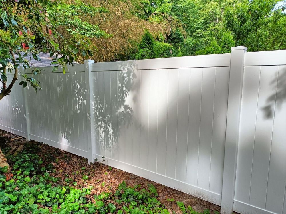 White vinyl privacy fence in a backyard, partially shaded by trees and foliage.