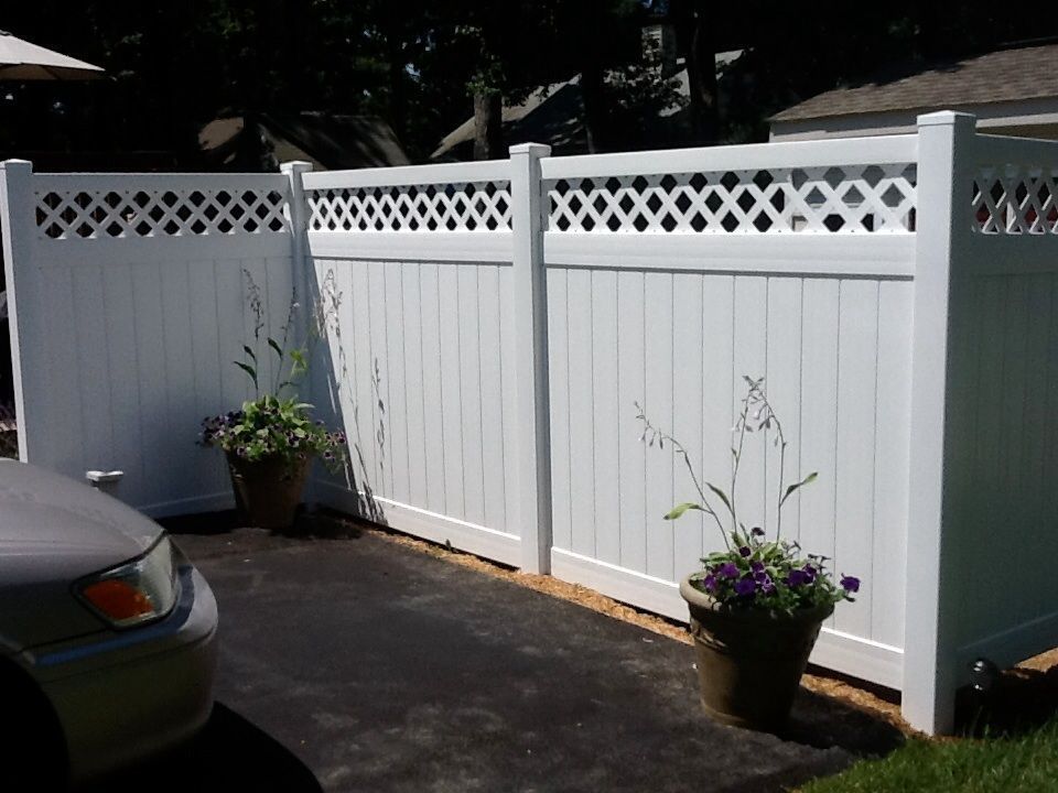 White vinyl fence with lattice top, potted flowers, and a car parked on blacktop.
