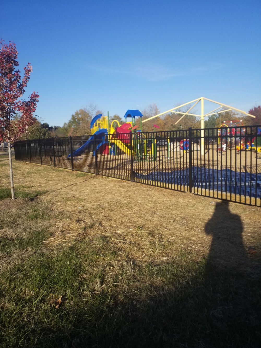 Black fence surrounds a playground with slides, swings, and shade structure, under a blue sky.