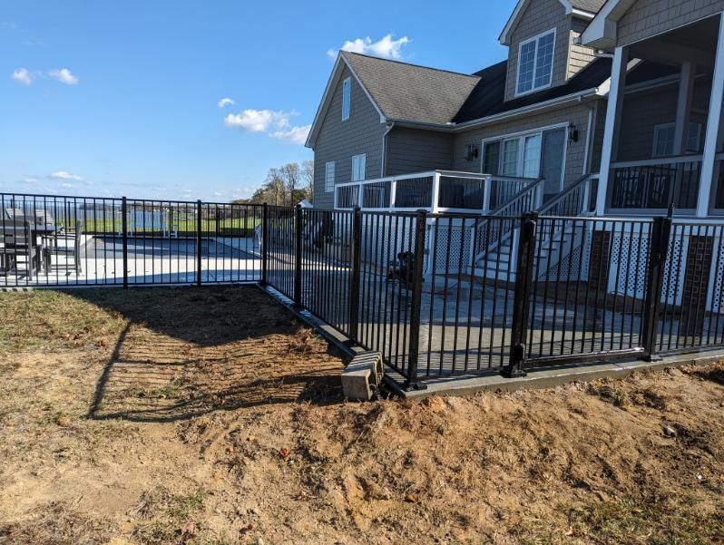 Black metal fence encloses a patio and pool area next to a light-colored house under a blue sky.