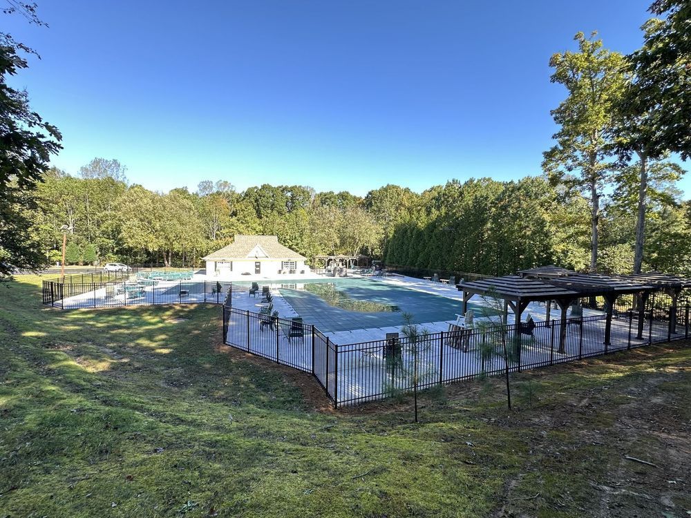 Outdoor pool area surrounded by a black fence and greenery on a sunny day. A building is visible in the background.