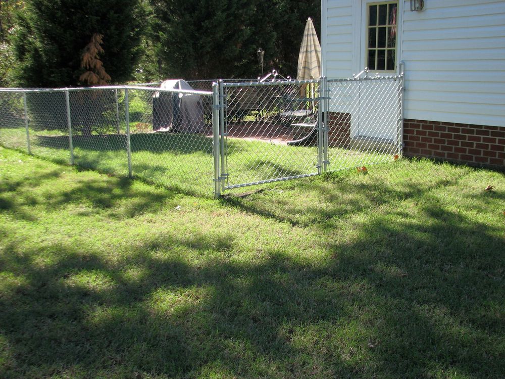 Chain link fence in a grassy yard, with a gate leading to a patio beside a white house.