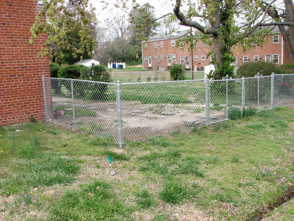 Chain-link fence encloses a dirt patch in a yard, next to a brick building.