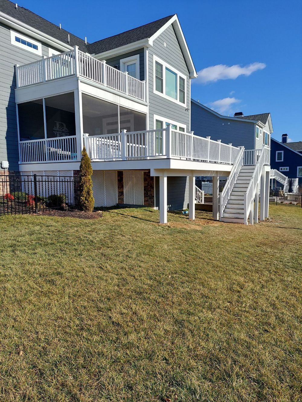 Two-story house with a white deck, stairs, and screened-in porch, over a grassy yard on a sunny day.