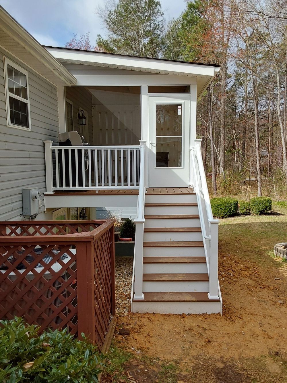 White screened porch with steps, attached to a gray house, set in a yard with trees.