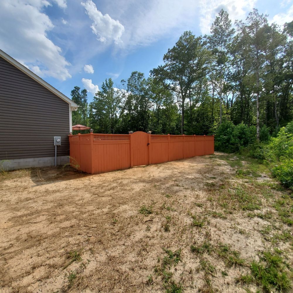 Orange fence encloses a backyard with dirt ground, adjacent to a house with brown siding and trees under a blue sky.