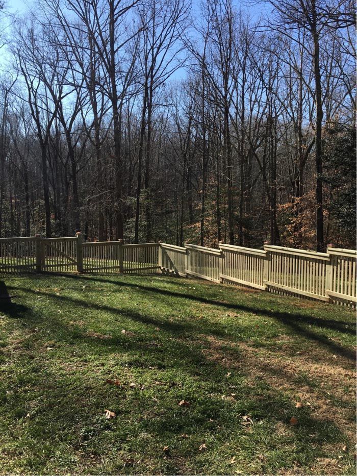 Wooden fence along the edge of a grassy yard with trees in the background under a blue sky.