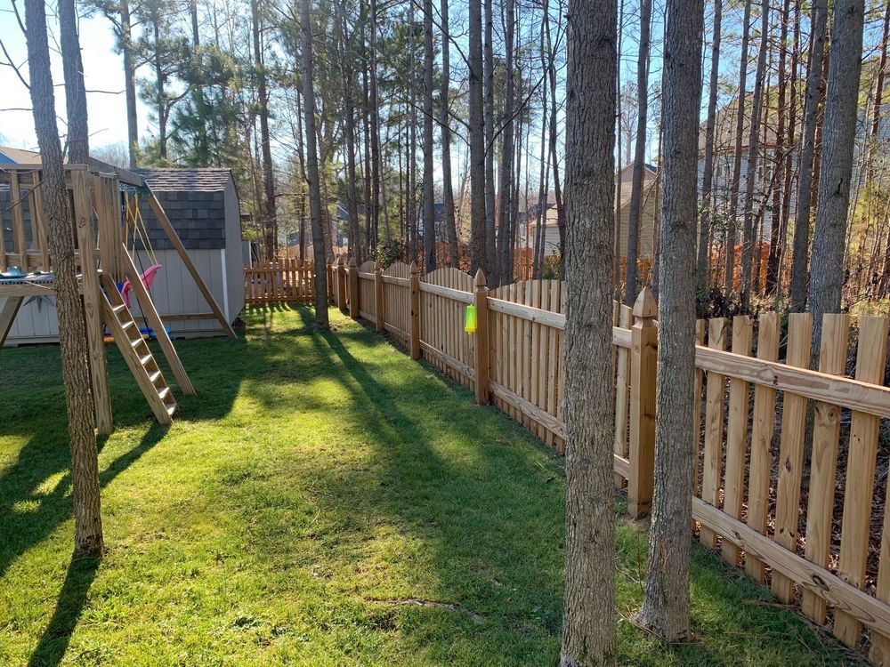 A wooden fence lines a backyard with green grass, trees, and a playset under a blue sky.