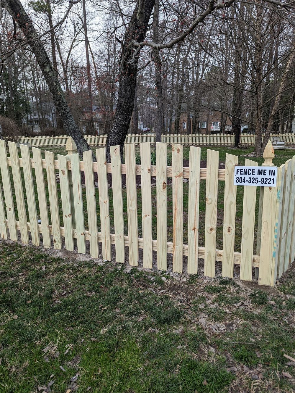 A light wooden picket fence surrounds a yard with a tree in it; sign states: 