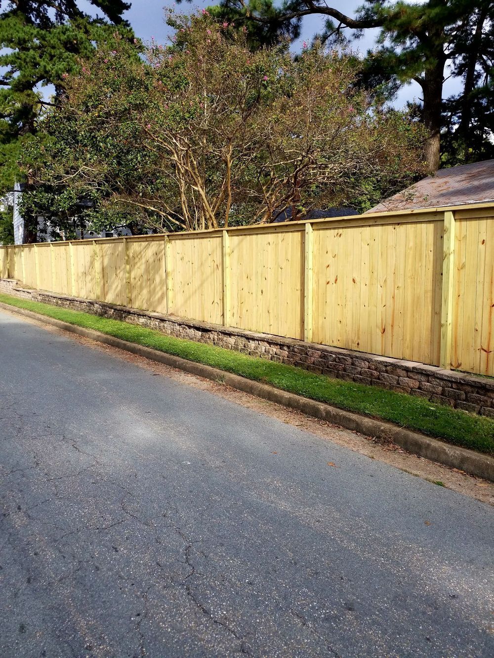 Wooden fence lines a street with green grass border, trees in the background.