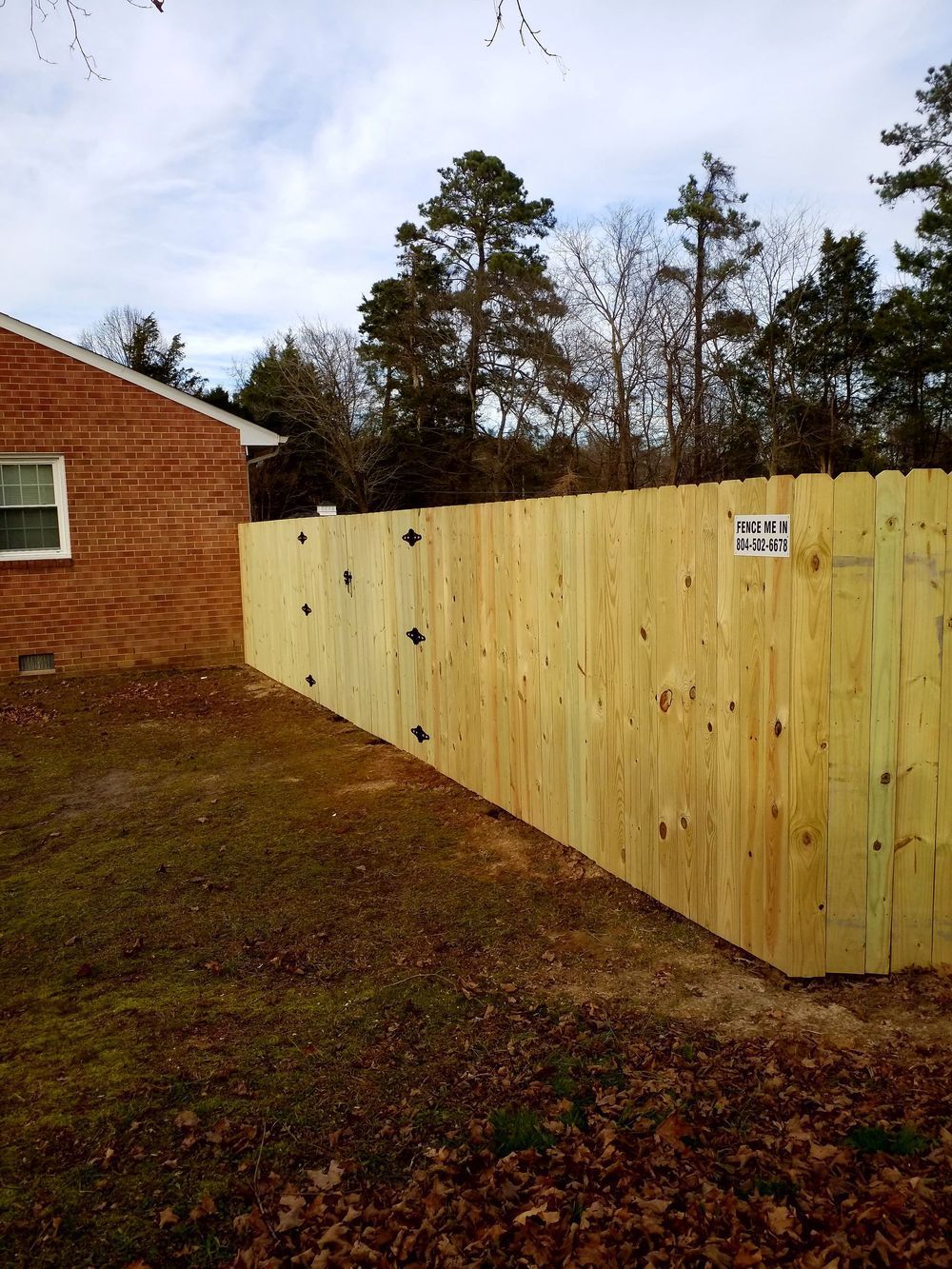 A new wooden fence built along the side of a brick house. Overcast sky and bare trees.