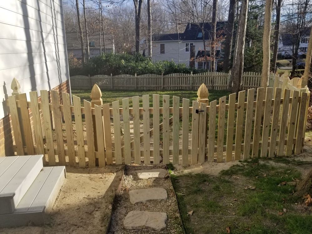 A wooden picket fence encloses a backyard with a gate, pathway, and trees in the background.