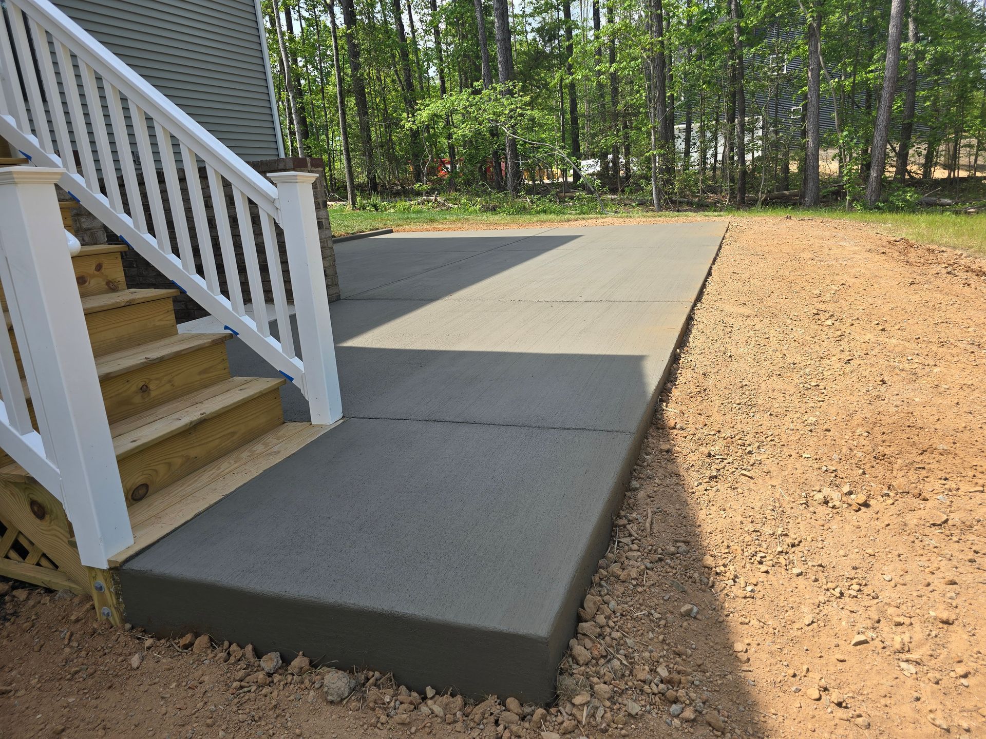 Concrete patio beside white stairs, bordered by mulch and surrounded by trees.
