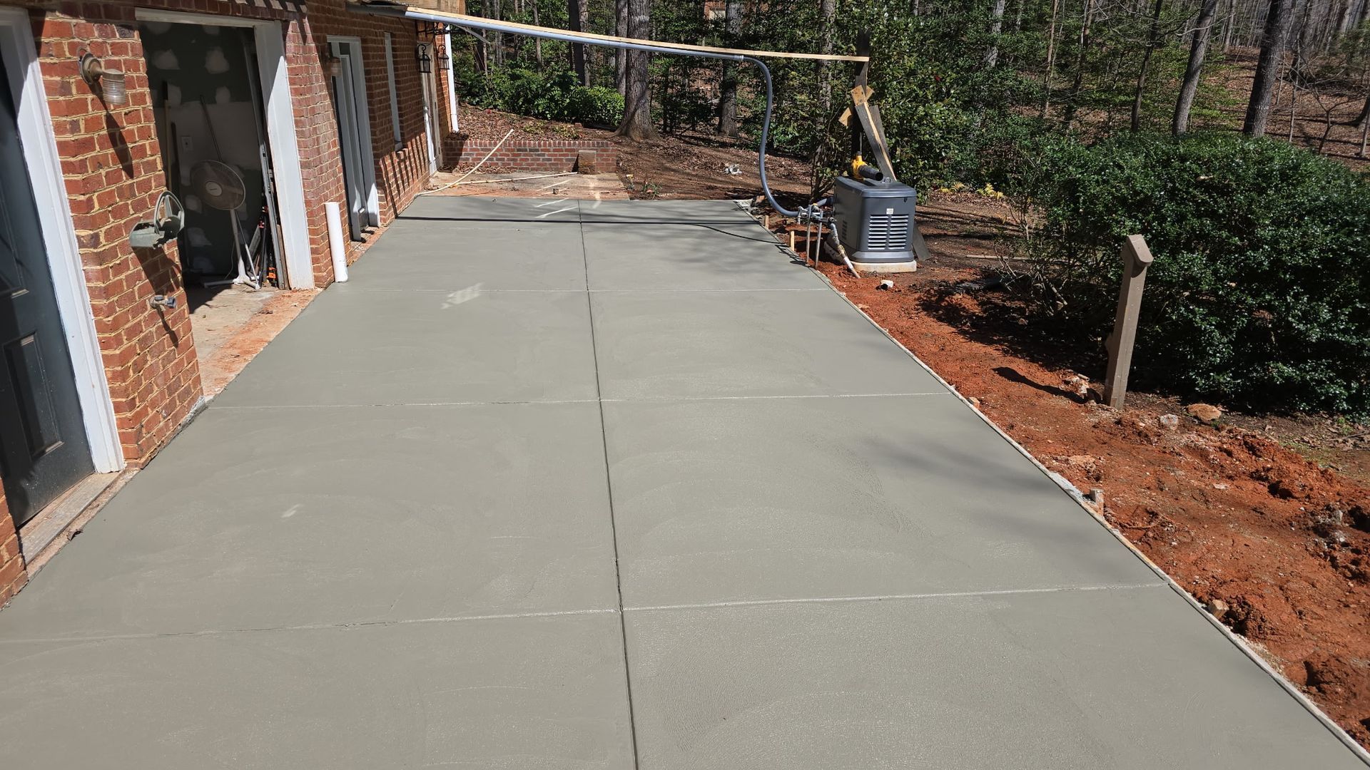 Freshly poured concrete walkway beside a brick house and dirt yard, leading toward a wooded area.