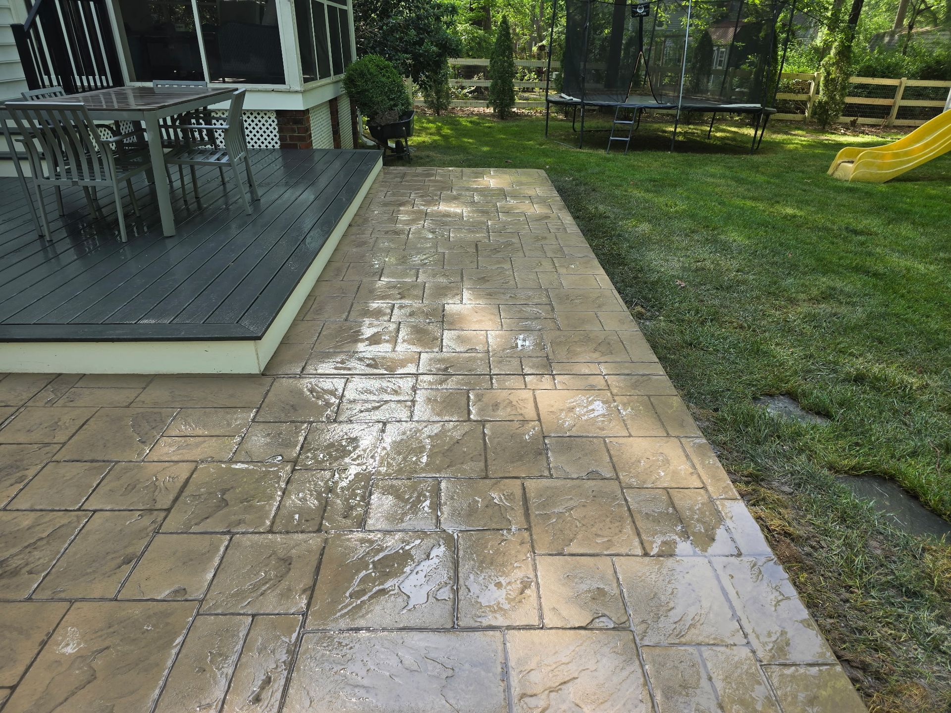 Wet stone patio beside a deck, with grass and a yellow slide in a backyard.