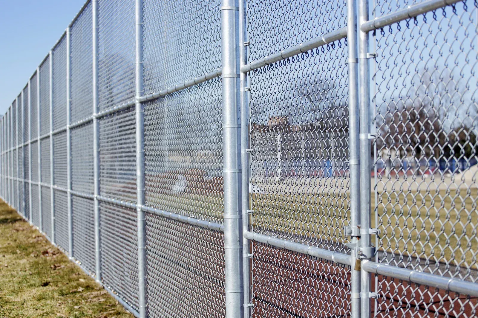 Chain-link fence in a grassy area, with a gate and a glimpse of a park in the background.