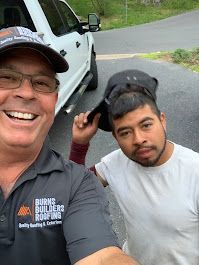 Two men smiling at camera in front of white truck. One holds a cap, other wears a branded shirt and cap.